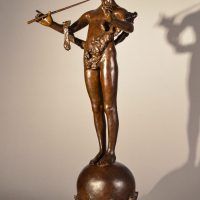 Alt text: Bronze fountain sculpture of the deity Pan mischeviously playing two flutes, standing atop a sphere flanked by fish, angled view