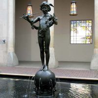 Alt text: Bronze fountain sculpture of the deity Pan playing two pipes and standing atop a sphere flanked by fish spouting water from the below pool