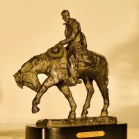 Alt text: Bronze sculpture of a cowboy riding on a horse with its front left leg outstretched and head down, angled view
