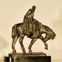 Alt text: Bronze sculpture of a cowboy riding on a horse with its front left leg outstretched and head down, side view