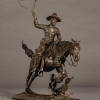 Alt text: Bronze sculpture of a cowboy with a lasso riding a horse jumping over a cactus, angled view