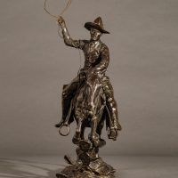 Alt text: Bronze sculpture of a cowboy with a lasso riding a horse jumping over a cactus, frontal view