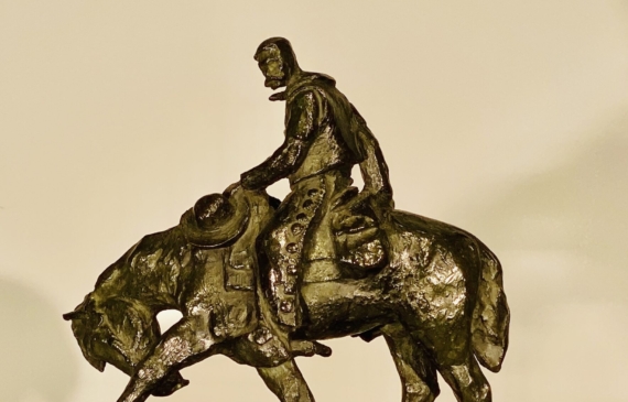 Alt text: Bronze sculpture of a cowboy riding on a horse with its front left leg outstretched and head down, side view